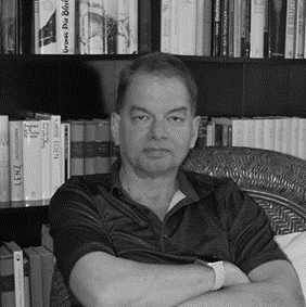 Black and white image of Chris Adami sitting with his arms crossed looking at the camera. He is sitting in front of a bookshelf full of books. 